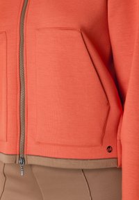 Korallfärgad zip-up hoodie med två framfickor, i en texturerad tyg och med kontrasterande brun kant. Utmärkande logotypdetalj på den nedre sidan.
