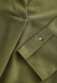 Olívovo zelená látková košela s hladkým povrchom, so skladaným rukávom, detailom gombíka a ostrým dizajnom švov.
