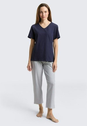 Femme debout pieds nus portant un haut bleu marine à manches courtes en V et un pantalon blanc à petits pois sur un fond clair.