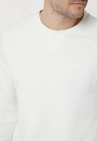 Felpa bianca con logo "GUESS" in rilievo al centro, colletto rotondo e maniche raglan, realizzata in morbido cotone.