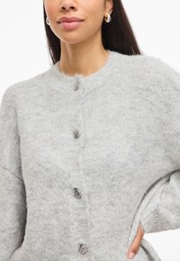 Pilkas megztas kardiganas su apvalia kaklo iškirpte, tekstūruotu audiniu, dideliais mygtukais ir laisvomis rankovėmis. Minkšta, jauki išvaizda su subtiliais pokyčiais.