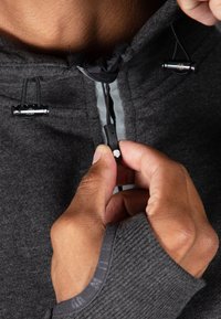 Felpa grigia con cappuccio e chiusura con cordino; presenta toggle elastici neri e un polsino strutturato. Focus sul meccanismo della zip e sul colletto.