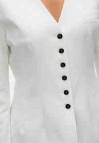 Blazer blanco con un escote en V profundo, que cuenta con seis botones negros en la parte delantera y un pequeño bolsillo en el lado izquierdo. Textura de tela suave.