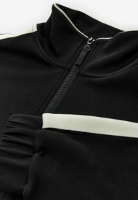 Chaqueta negra con cremallera y cuello alto, con franjas de acento blancas, textura suave y cierre central de cremallera.