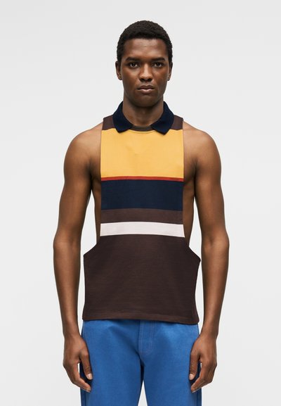 Άνδρας φοράει αμάνικη, πολύχρωμη μπλούζα πόλο με κίτρινες, ναυτικές, καφέ και λευκές ρίγες, και μπλε παντελόνια, στέκεται μπροστά σε απλό φόντο.