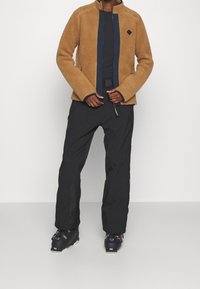 Brązowa polarowa kurtka z wysokim kołnierzem i granatowym zamkiem, zestawiona z czarnymi wodoodpornymi spodniami i butami narciarskimi. Teksturowany, ciepły materiał.