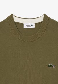 Maglione a maglia verde oliva con colletto rotondo, caratterizzato da un bordo bianco strutturato e un piccolo logo del coccodrillo ricamato sul lato sinistro del petto.