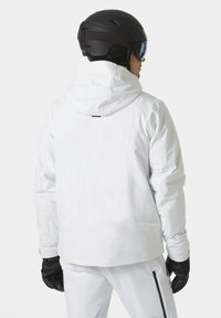 Bílá lyžařská bunda s kapucí, strukturovaná tkanina a kapsy na zip. Nositá s černými rukavicemi a helmou, ukazující elegantní zadní design.