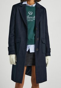 Una persona indossa un cappotto scuro sopra un felpa verde e una camicia a righe blu, con una gonna grigia e guanti lavorati a maglia, in piedi su uno sfondo chiaro.