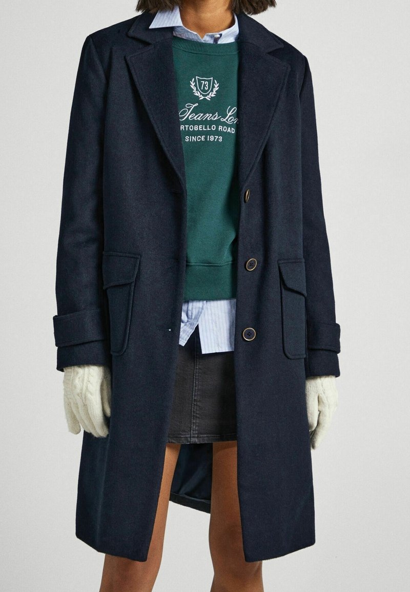 Una persona indossa un cappotto scuro sopra un felpa verde e una camicia a righe blu, con una gonna grigia e guanti lavorati a maglia, in piedi su uno sfondo chiaro.
