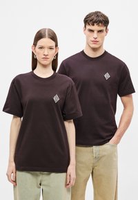 Ciemnobrązowe bawełniane koszulki z białym logo w kształcie diamentu na piersi. Obaj modele mają na sobie spodnie dżinsowe o luźnym kroju.