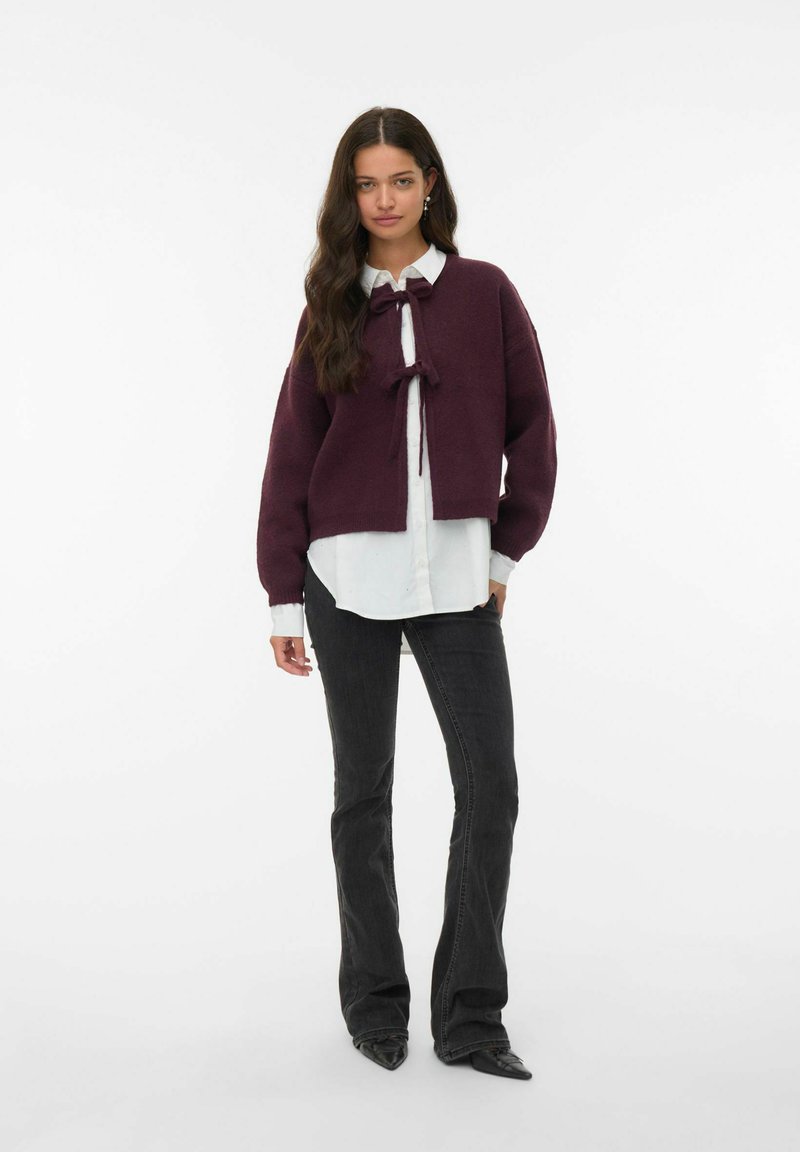 Pull cropped bourgogne avec un nœud à l'avant, superposé à une chemise blanche à boutons. Assorti avec un jean noir évasé et des chaussures noires pointues.