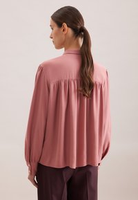 Blusa a maniche lunghe color rosa pallido in tessuto morbido con dettaglio a piega sul retro, polsini con bottone e colletto morbido. Indossata con pantaloni scuri.