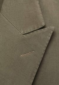Detalj maslinasto zelene tkanine jakne koji prikazuje teksturirano tkanje, izrezani rever i šavove. Sadrži mali naglasak blizu revera.