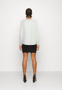 Weiße Bluse mit langen Ärmeln und schwarzen Punkte, kombiniert mit einem eng anliegenden schwarzen Rock. Die Bluse hat einen lockeren Schnitt und knöpfbare Manschetten.