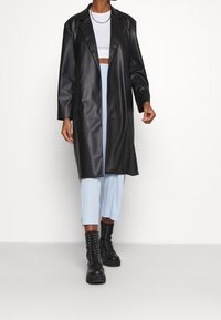 Person som bär en lång kappa i svart konstläder, en vit crop top, ljusblå korta byxor, svarta kängor och ett silverkedjehalsband.