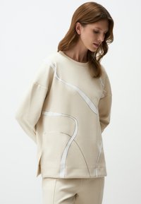 Beige sweatshirt met lange mouwen, voorzien van abstracte witte lijnpatronen. Zacht fabric, ontspannen pasvorm en zijsplitten aan de onderkant.