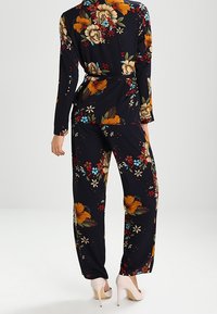 Tuta floreale con sfondo nero, caratterizzata da grandi fiori arancioni, gialli e blu. Maniche lunghe, vita con cintura, pantaloni a gamba larga.