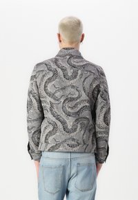 Grå jakke med slange mønster, struktureret stof og en skræddersyet pasform, parret med lyseblå jeans, vist fra ryggen.
