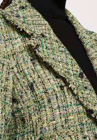 Gros plan d'une veste en tweed vert texturé avec des fils orange et noirs portée sur un col roulé côtelé noir.