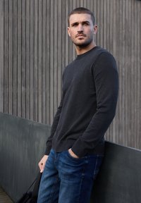 Homme aux cheveux courts portant un pull sombre et un jean bleu, debout à l'extérieur, une main dans la poche et tenant un sac de l'autre.