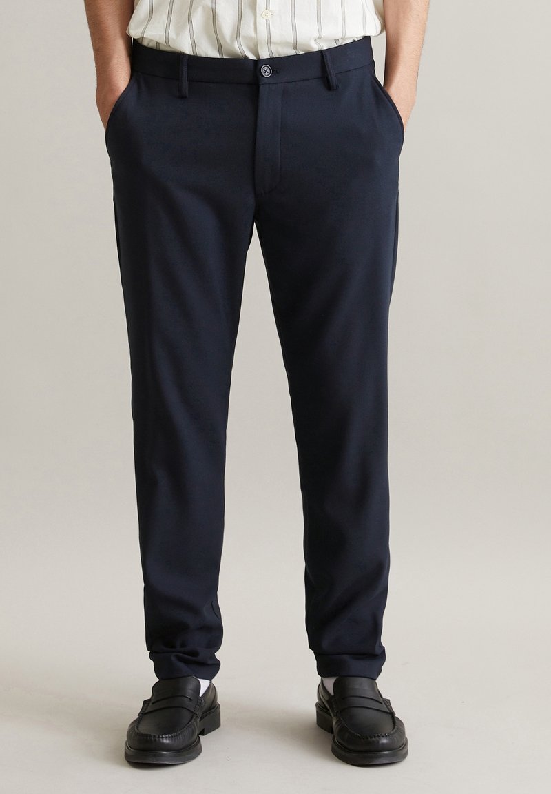 Homme portant un pantalon bleu marine foncé sur mesure, des mocassins noirs, des chaussettes blanches et une chemise blanche à fines rayures verticales rentrée à l’intérieur. Mains dans les poches.