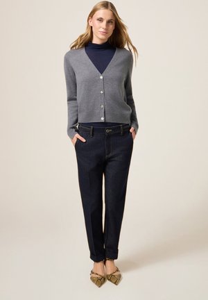 Graue Kaschmirstrickjacke mit V-Ausschnitt und Knöpfen, getragen über einem marineblauen Rollkragenpullover, kombiniert mit dunklen Denim-Hosen und flachen Schuhen im Schlangenmuster.