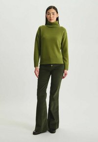 Pull à col roulé tricoté vert avec un ourlet côtelé, associé à un pantalon en velours côtelé vert foncé évasé. Le modèle se tient devant un fond neutre.