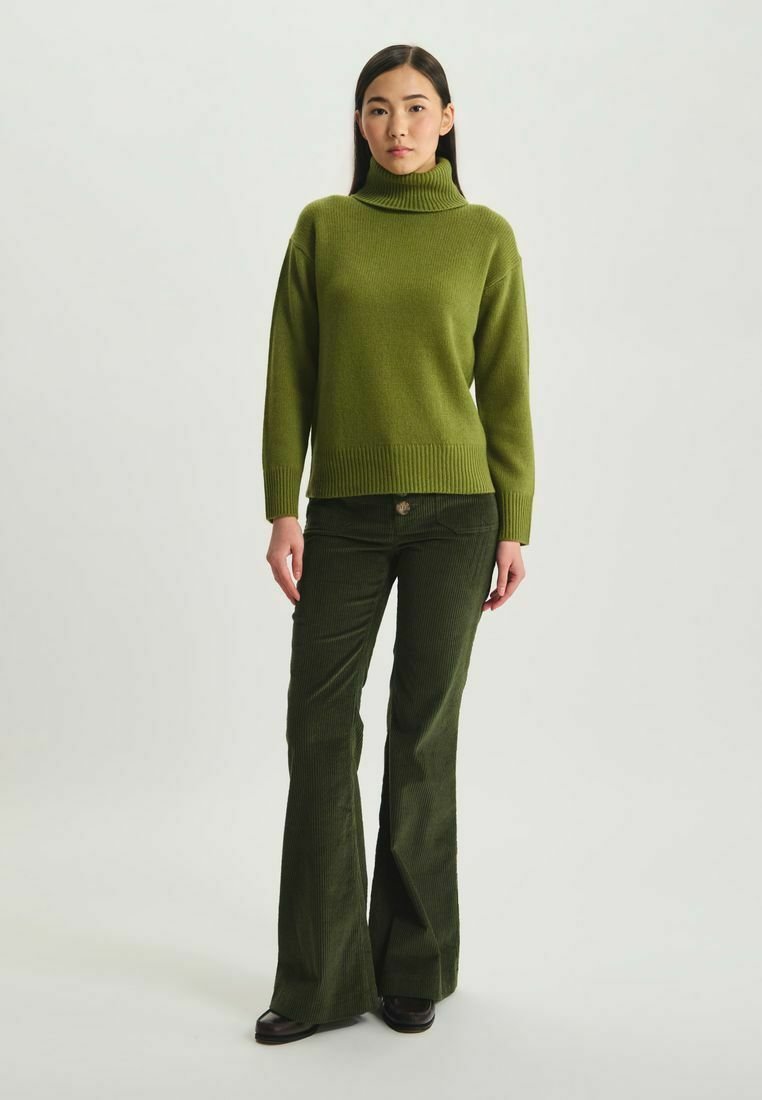 Pull à col roulé tricoté vert avec un ourlet côtelé, associé à un pantalon en velours côtelé vert foncé évasé. Le modèle se tient devant un fond neutre.