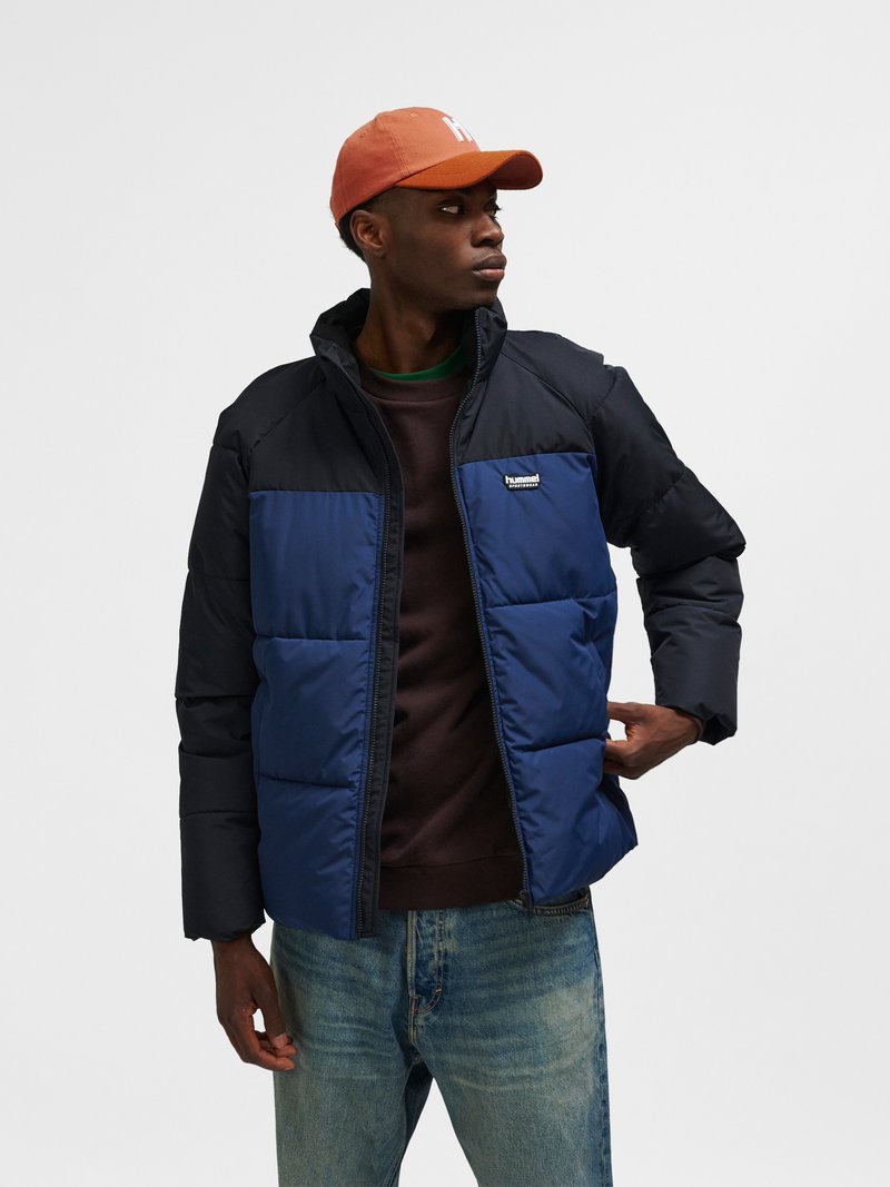 Homme portant une doudoune bicolore bleu marine et noire, une casquette orange, un sweat-shirt foncé et un jean bleu, regardant vers la droite avec la main dans la poche.
