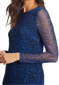 Žena v modro-černém vzorovaném topu s dlouhými rukávy z jemného síťového materiálu s vířícími abstraktními vzory, s rukama jemně spojenýma u pasu.