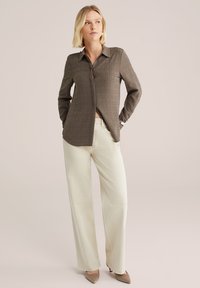 Modèle portant une chemise à boutons texturée marron et un pantalon large crème, posé devant un fond neutre, dégageant une allure stylée et décontractée.
