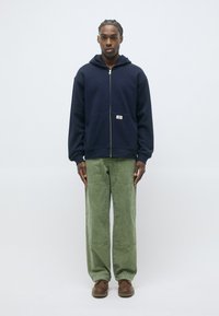 Felpa con zip di colore blu navy, con un ampio cappuccio e tasca frontale, abbinata a pantaloni a gamba larga verdi e scarpe marroni. È visibile una texture di tessuto liscia.