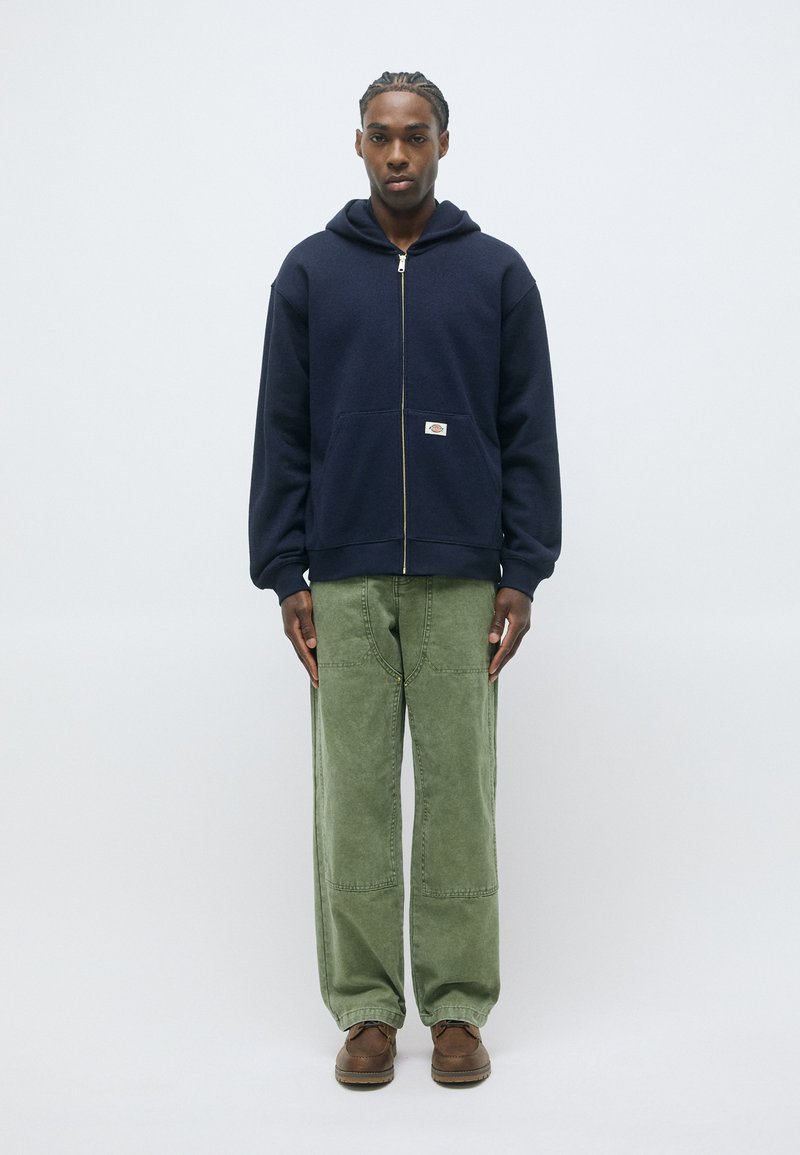 Felpa con zip di colore blu navy, con un ampio cappuccio e tasca frontale, abbinata a pantaloni a gamba larga verdi e scarpe marroni. È visibile una texture di tessuto liscia.