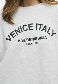 Siva dukserica s crnim natpisom "VENECIJA ITALIJA LA SERENISSIMA EST.421AD", s okruglim izrezom i mekim materijalom.
