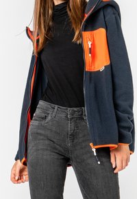 Fleece takki tummansinisellä värillä oransseilla yksityiskohdilla, jossa on vetoketjullinen rintatasku, huppu ja elastiset hihansuut. Yhdistetty mustiin farkkuihin.