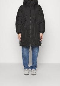 Černá péřová bunda s kapucí, předním zipem a bočními kapsami. Má volný střih a elastické manžety. Kombinováno se světle modrými džíny a šedými teniskami.