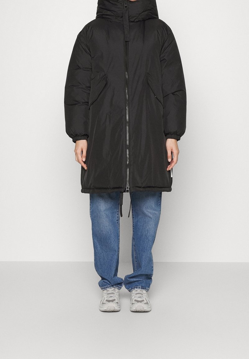 Černá péřová bunda s kapucí, předním zipem a bočními kapsami. Má volný střih a elastické man�žety. Kombinováno se světle modrými džíny a šedými teniskami.