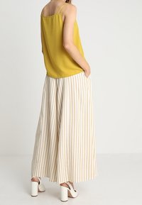 Ärmlös senapsgul topp med tunna axelband, kombinerad med vida, elfenbensfärgade byxor med vertikala beige ränder. Modell bär vita klackskor.