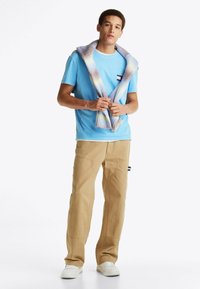 Camiseta de algodón azul claro con cuello redondo blanco, pantalones cargo beige y un suéter de punto multicolor sobre los hombros. Zapatillas blancas.