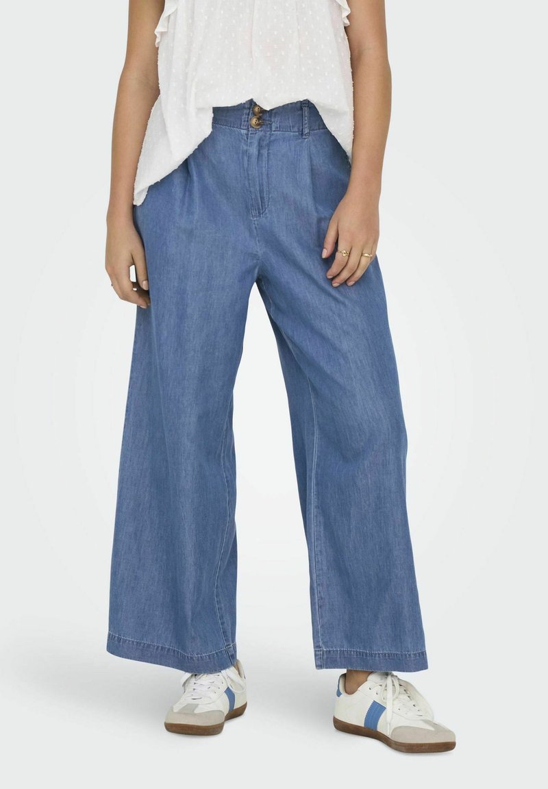 Pantalones de mezclilla anchos de color azul claro con una cintura alta, dos bolsillos frontales y cierre de botón, combinados con zapatillas blancas.