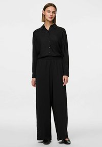 Črna srajca s dolgimi rokavi in prsno žepom, v kombinaciji z visokimi črnimi širokimi hlačami; gladka tkanina in sproščen kroj.