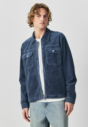 Fiatal férfi hullámos szőke hajjal, kék kordbársony dzsekit visel fehér ing fölött és világoskék farmert, sima szürke háttér előtt áll.