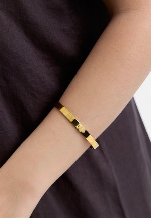 Złota metalowa bransoletka w formie bangla z gładką, wypolerowaną powierzchnią i małym dekoracyjnym emblematem. Noszona na nadgarstku na tle ciemnej tkaniny.