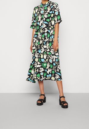 Femme portant une robe midi noire ornée de formes et de symboles abstraits colorés, associée à des sandales noires à talons épais.