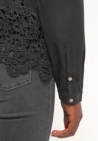 Haut noir à manches longues avec des détails en dentelle, associé à un jean en denim gris foncé, montrant une main reposant à côté de la hanche.
