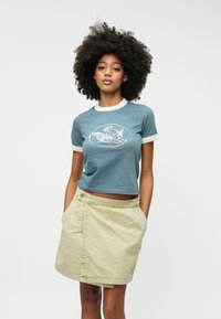 Bomulds croppede t-shirt i teal med creme ribbet halsudskæring og ærmer, der har et centralt hvidt grafisk motiv. Sammen med en beige denimskørt.