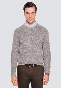 Muž v šedém svetr s texturou přes světle modrou košili s límečkem, hnědých kalhotách a hodinkách, stojící před jednobarevným světle šedým pozadím.