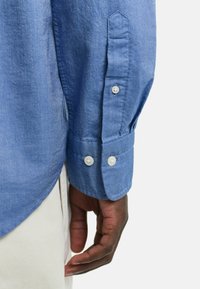 Lys blå, knappet skjorte laget av et teksturert stoff. Har krage med spisser og knapper på mansjettene, med synlige sømdetaljer.