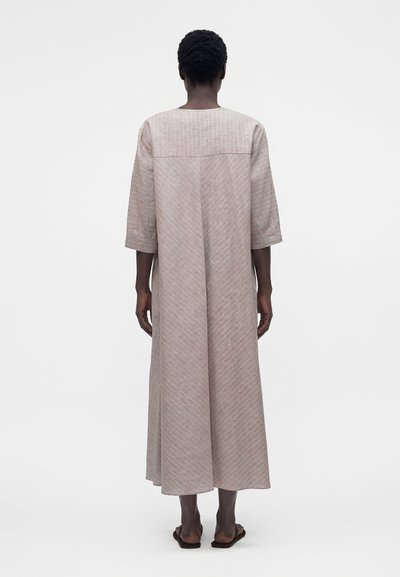 Άτομο με κοντά σγουρά μαλλιά, φοράει ένα φαρδύ, μακρύ, μπεζ φόρεμα με σχέδιο και μανίκια τριών τετάρτων, όρθιο, ξυπόλυτο, με λευκό φόντο.